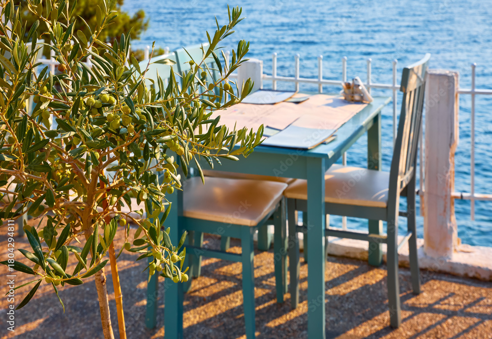 Obraz premium Sithonia, Chalkidiki, Greece. Traditional tavern cafe exterior with wooden table chairs. Olive tree branch focused in front with the view of the aegean sea in the background..