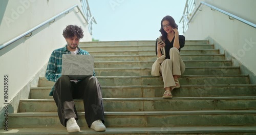 Indian young adult gen z busy male man use laptop look screen do study check online e work at campus place. Happy female sitting on stair hold mobile phone read notes enjoy new day spend break time