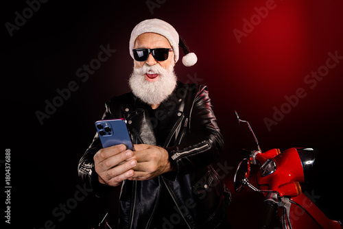 Cool Santa rocker with leather jacket posing on a red scooter using a phone during Christmas festive mood