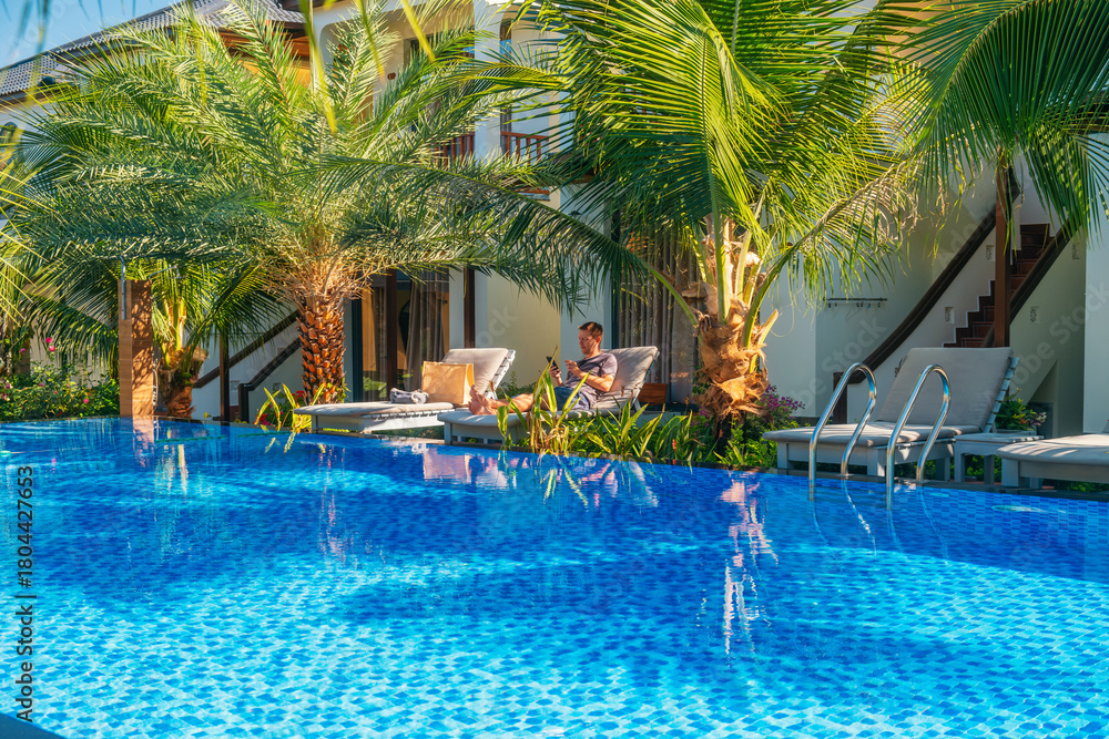 Obraz premium Man with smartphone relaxing on sun lounger by the swimming pool at luxury hotel surrounded by palm trees on Phu Quoc island, Vietnam. Tourist resting in topical resort on summer vacation in Asia