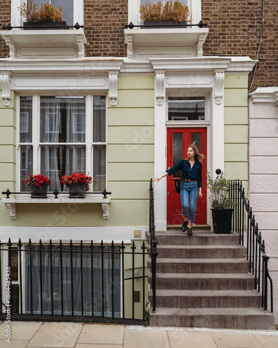 A young woman descends the concrete steps of a charming light green house with a vibrant red door in London Borough of Camden. Her hair gently blows in the breeze.