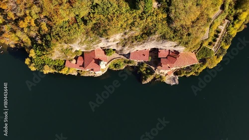 Drone flyover of Santa Caterina del Sasso carved into the cliff above Lake Maggiore, its terracotta roof glowing among autumn colors in the Piedmont landscape.