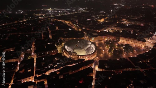 Majestic evening aerial of Verona with the illuminated Arena di Verona amphitheatre of 12 century, glowing city lights and historic streets creating a cinematic night atmosphere