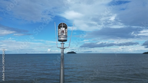 Close-up of a metallic navigation light on a pier with a vast blue sea and cloudy sky in the background.