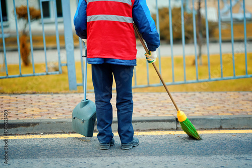 Municipal street cleaner in red vest working outdoors, sweeping road and sidewalk as part of everyday public sanitation and urban maintenance duties. Utility employee sweeping road