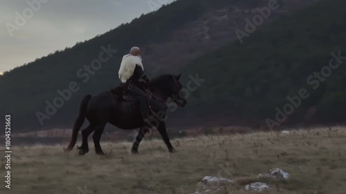 Cinematic aerial drone shot of a Viking warrior on a large black horse surveys a dramatic landscape during sunset, wearing a fur cloak concept of Ancient exploration, adventure, and the spirit of the