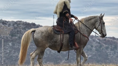 Aerial drone establishing or tracking shot of Bearded warrior in fur cloak riding a white horse across open landscape at sunrise, mountains in the background. Historical, rugged, adventurous scene