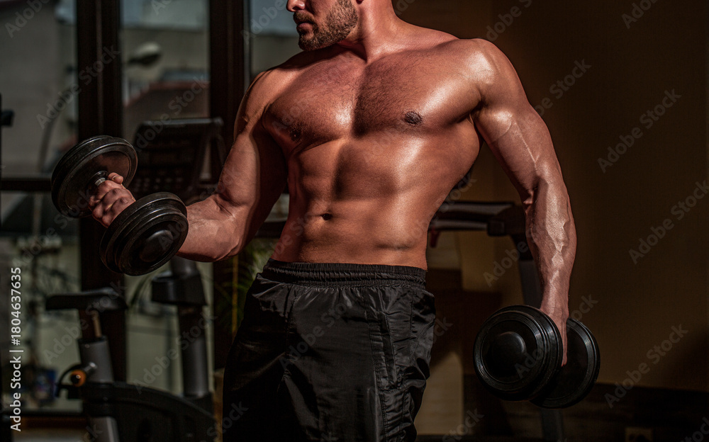 Naklejka premium Man lifting dumbbell in a gym making exercise for muscles, dumbbells. Power athletic man in training pumping up muscles with dumbbell. Strong bodybuilder with perfect biceps curl, triceps and chest