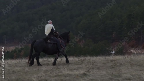 Cinematic aerial drone shot of a Viking warrior on a large black horse surveys a dramatic landscape during sunset, wearing a fur cloak concept of Ancient exploration, adventure, and the spirit of the