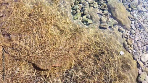 Clear water flowing over rocky shoreline with sunlit reflections and pebbles