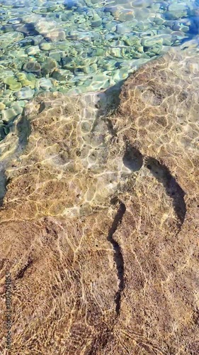 Sunlit coastal waters flow over rocky shoreline with shimmering waves and crystal clarity