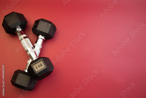 Black dumbbells, each weighing 2 kg, are positioned in a crossed formation on a bright red background, highlighting fitness and strength training essentials