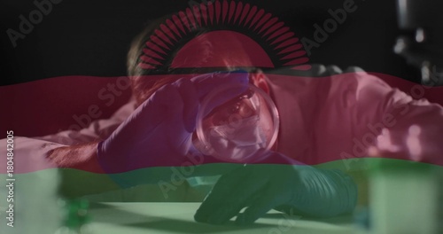 Leaning scientist holding glass sphere to eye at lab bench, white coat, blue gloves, flag overlay