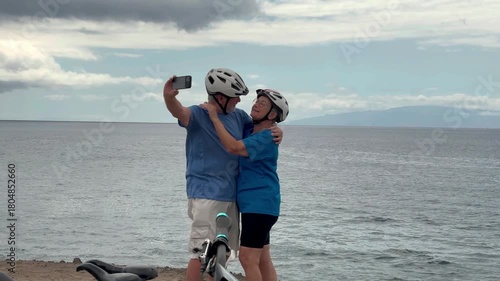 Happy senior couple of cyclists wearing helmets exchange a kiss and take a selfie with phone enjoying riding seaside with electric bicycles. Healthy lifestyle in retirement. Horizon over the sea