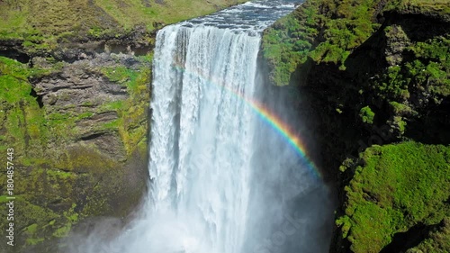 Aerial view of Skogafoss waterfall from a side angle. Drone footage showing dense mist rising from the falls and a rainbow appearing in the sunlight.