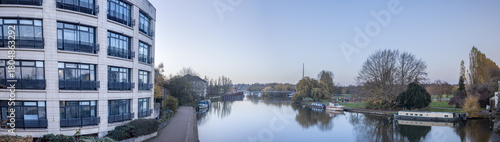 Panoramic daylight view of a river with houseboats and parkland on the far bank, framed on the left by the cream-colored facade of a curved building.