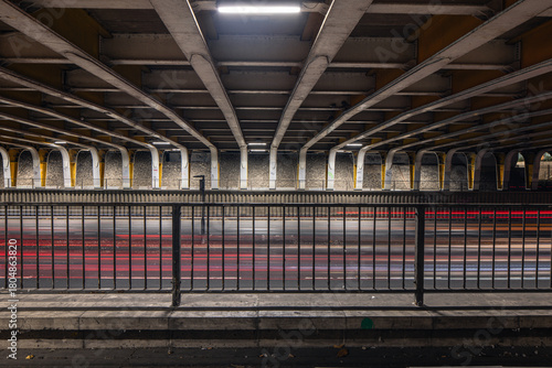 A symmetric view of a busy roadway under a bridge, featuring numerous repeating structural beams and red light trails from traffic, separated by a black metal railing.