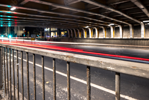 A low-angle, long-exposure photograph from a pedestrian area beneath an overpass, highlighting a weathered metal railing and the streaking red and white lights of traffic.