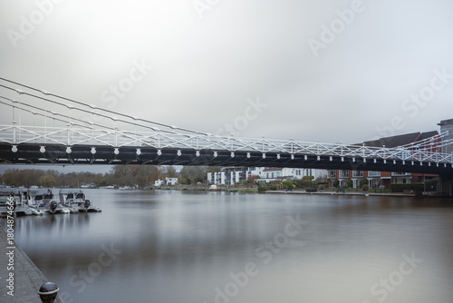 Photograph taken with a long exposure of a suspension bridge crossing a calm river, with the water's surface smoothed out. Boats are moored to the left, and houses line the opposite bank.