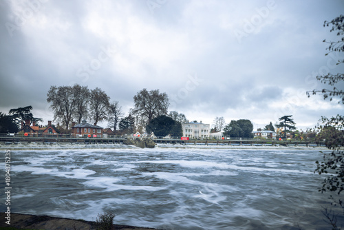 A wide, long-exposure photograph of a river flowing over a weir, with white-capped rapids in the foreground, and residential buildings and trees visible on the far bank under an overcast sky.