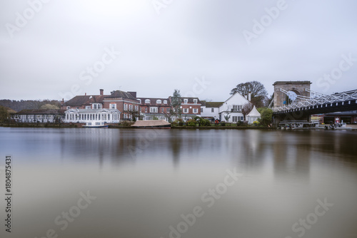 A long-exposure photograph across a very smooth, reflective river showing a row of grand, traditional buildings, including a large white structure and red-brick houses , marlow