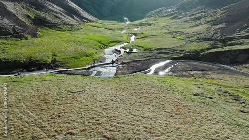 Drone view of Reykjadalur geothermal river in bright green valley in Iceland. Drone view of tourists walking near the hot spring river as it winds through a mossy mountain landscape.