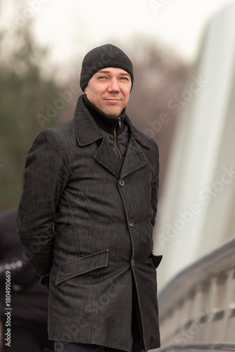 40-year-old man walking in a hat and trench coat. Smiling middle-aged man. Men's autumn fashion. View from the back. Look into the camera. Walking alone on a city street. Good face. Three-day stubble.