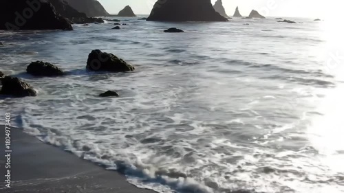 Ocean waves washing over a sandy shore with large rock formations in the background at sunrise.