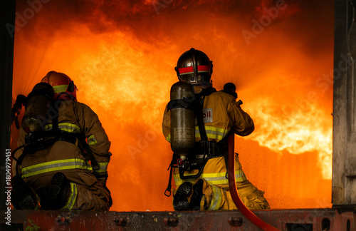 Two firefighters in full protective gear work together to extinguish a massive blaze