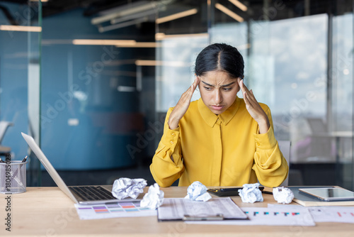 A sad young Indian woman sits frustrated at an office desk with crumpled paper and holds her head in frustration