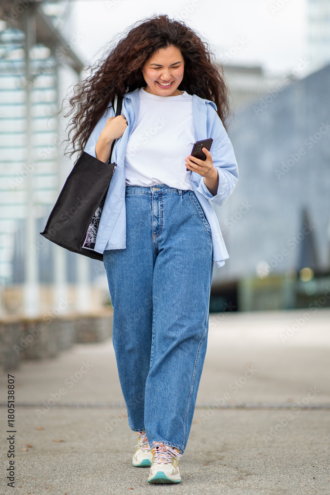 Fototapeta premium Cheerful young Asian woman walking while smiling at smartphone on city street