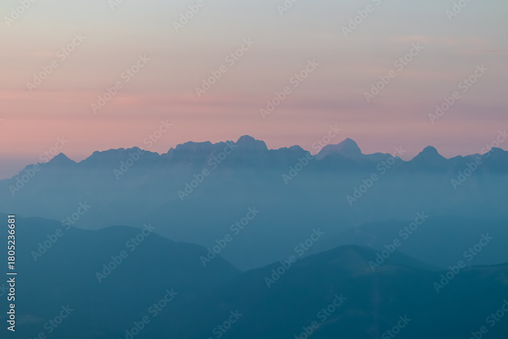 Fototapeta premium The majestic peaks of a distant alpine range emerge through layers of blue morning mist, viewed from Dobratsch. A serene and moody mountain landscape captured under a soft, pastel-colored dawn sky.