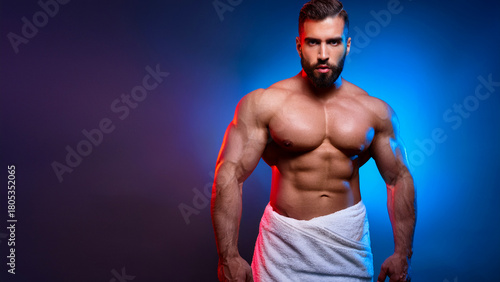 Muscular handsome man with dark beard poses shirtless in studio. Wears white towel, showing strong fit body with six pack abs, powerful chest.