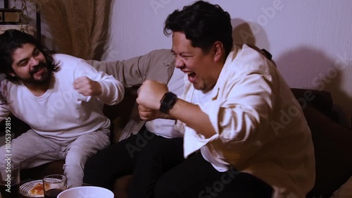 three young Hispanic men on a couch. They watch the screen intently, then cheer and raise their fists in shared celebration of a victory or successful outcome