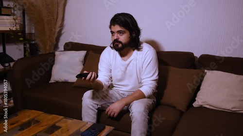 Close-up video of a young Hispanic man with long hair and a beard sitting on a couch, holding a remote control and changing channels, smiling while enjoying home entertainment