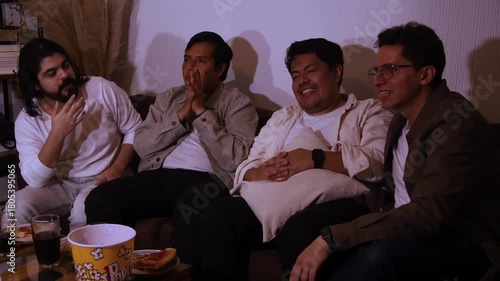 young Hispanic men on a couch, laughing and conversing while enjoying a show or movie. Highlights strong shared humor, social interaction, and close male bonding.