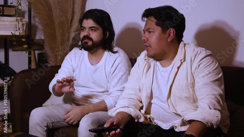 Hispanic men with long hair and a beard sitting on a couch, holding a remote control and laughing heartily. Highlights joyful home entertainment and media enjoyment.