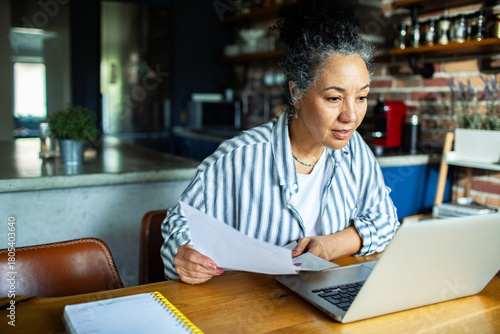 Mature woman reviewing finances on laptop at home kitchen, focused