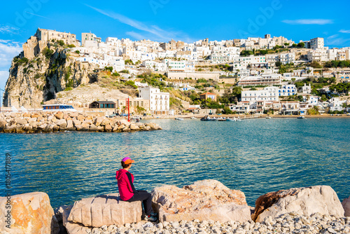 Female traveler sitting on coastal rocks admiring the whitewashed town of Peschici overlooking the sea, Apulia, Italy.