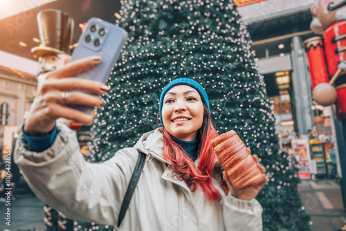 Happy young woman taking selfie with a smartphone and authentic Trdelnik at snowy European Christmas market