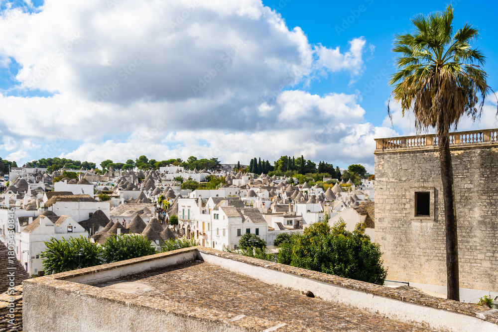 Naklejka premium Panoramic view of the trulli rooftops and palm trees under bright clouds and blue sky in Alberobello town, Apulia, Italy