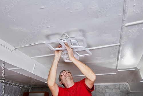 LED chandelier is positioned on ceiling while electrician performs installation in rural house connecting wires and securing fixture.