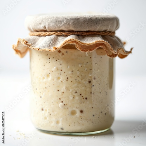 Glass jar filled with active bubbly sourdough starter culture. Starter is a beige fermenting mixture with visible air pockets and dark specks. It is covered with cloth and twine tied around the neck.