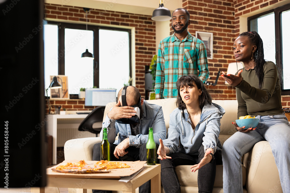Fototapeta premium Diverse friends gather at home, reacting to dramatic video game moment. Multiethnic men and women show disappointment while sitting on couch, holding VR headset and controllers during weekend hangout.