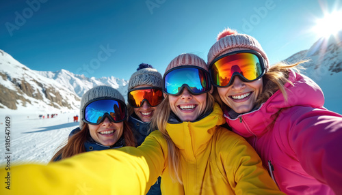 Four happy women friends take selfie on sunny winter day. Wear colorful ski jackets, bright goggles, warm hats. Group enjoys skiing vacation at snowy mountain resort, smiling together during fun trip.