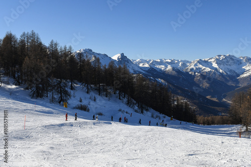 Skiers and snowboarders descending a ski slope in Sauze D'Oulx ski resort, Turin, Italy. Tree lined piste overlooking beautiful snowcapped mountains and the Milky Way ski area  in the Piedmont region.