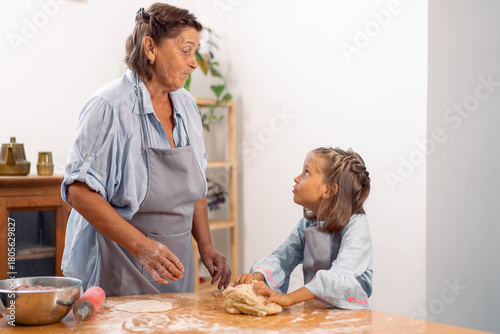 Girl and grandmother in aprons working together kneading dough on floured kitchen table enjoying family time, baking and learning at home