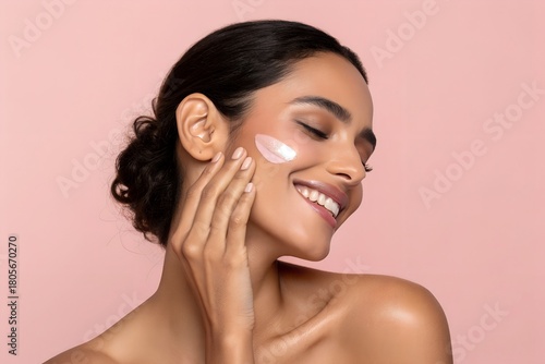 A smiling young woman gently applies white beauty cream to her glowing cheek, enjoying a fresh skincare routine with closed eyes.