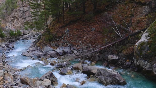 A scenic view of a fast flowing mountain river cutting through a rocky landscape in the North Caucasus region of Russia. The banks are covered with autumn foliage and fallen trees, highlighting the wi