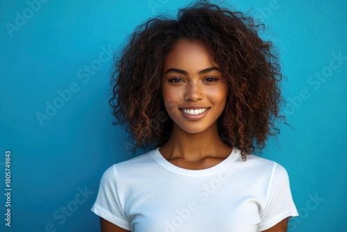 young person with voluminous curly hair in a white t-shirt standing calmly against a vibrant blue wall, relaxed neutral pose head-and-shoulders portrait